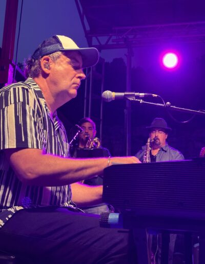 A keyboard player under purple stage lights with a horn section behind